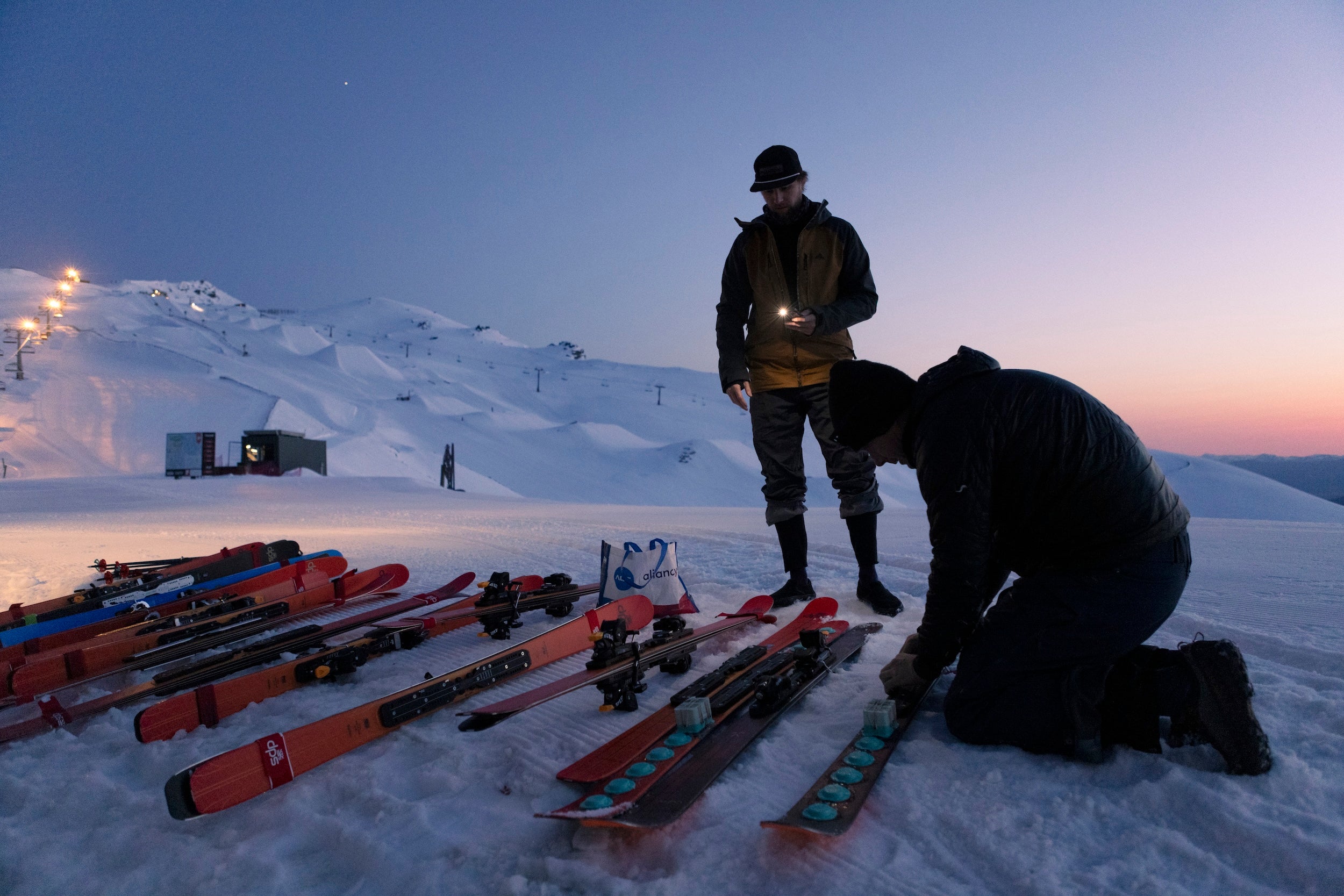 Two people preparing skis on a snowy mountain at dusk.